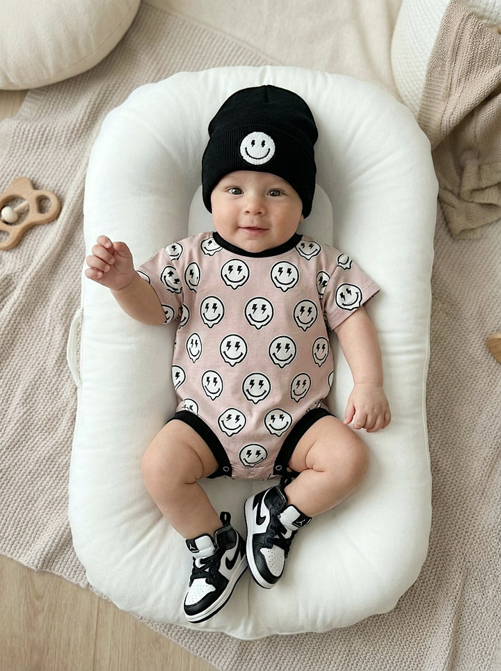 Baby wearing a smiley face onesie and hat, laying on a white cushion.