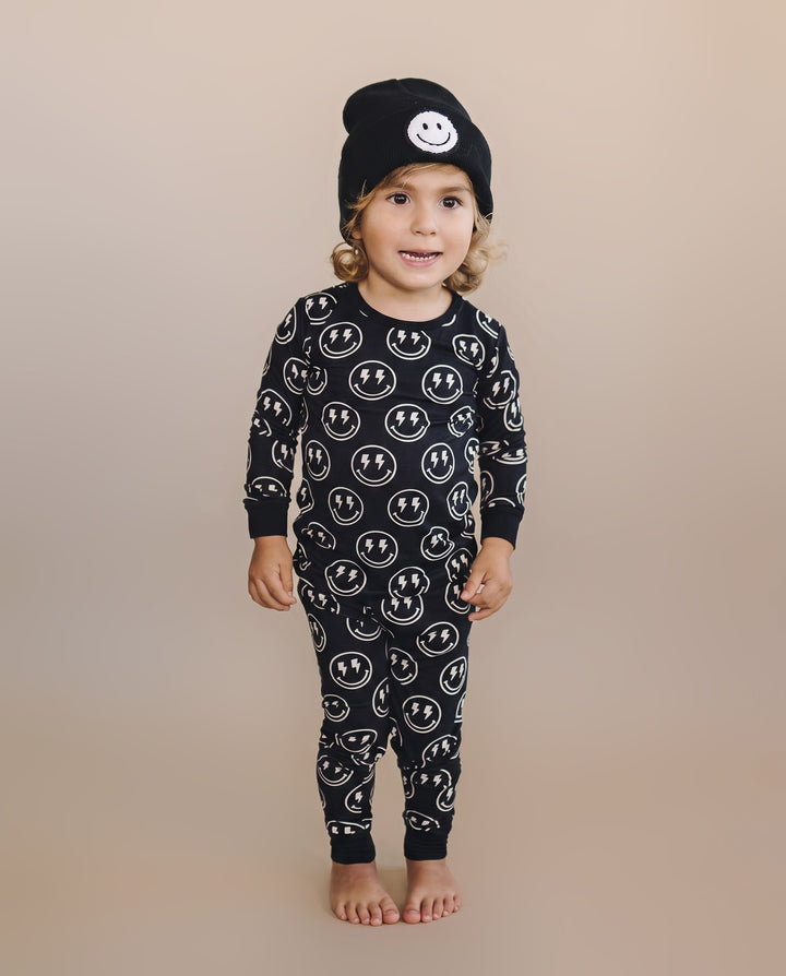 A toddler standing barefoot against a plain beige studio background, wearing a black long-sleeve one-piece pajama with an allover white smiley face print and a black beanie with a white smiley face patch on the front.