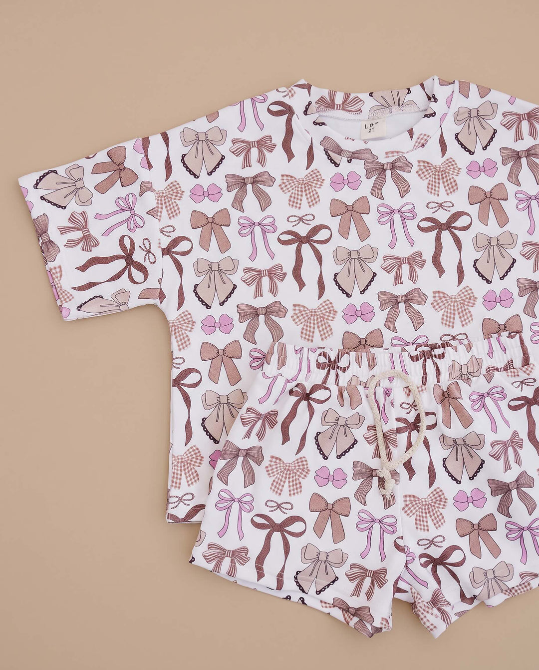 A two-piece baby romper and shorts set featuring an all-over bow pattern in shades of brown, mauve, and pink on a white background, laid flat on a beige background.