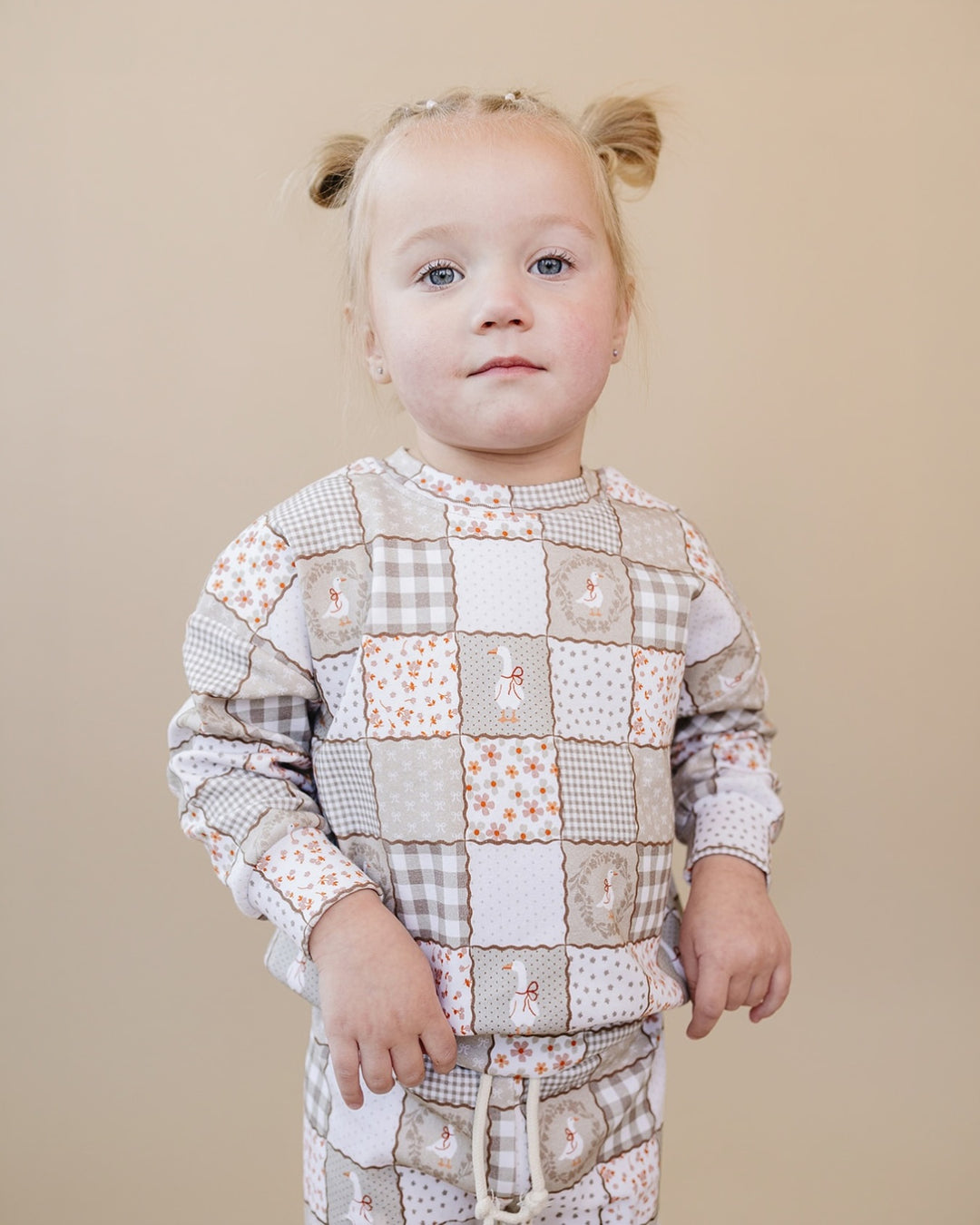 A toddler girl with blonde pigtails wearing a beige and white patchwork pajama set with gingham, floral, and bunny-print squares, standing against a solid beige background.