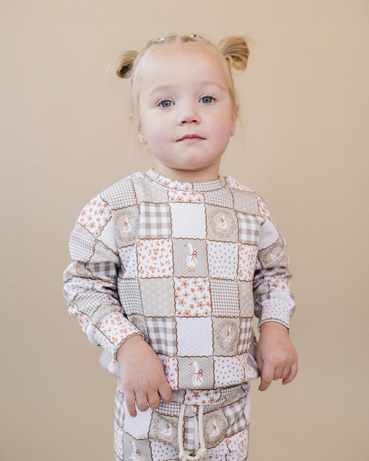 A toddler girl with blonde pigtails wearing a beige and white patchwork pajama set with gingham, floral, and bunny-print squares, standing against a solid beige background.