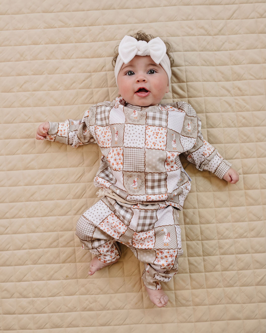 A baby lying on a beige quilted blanket, wearing a long-sleeve patchwork pajama set in beige, brown, and white with a white bow headband.