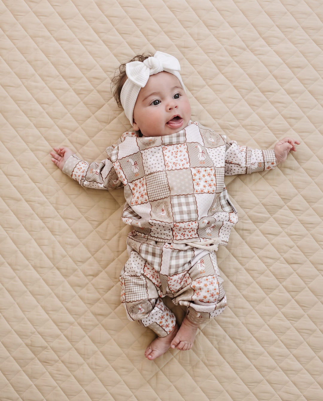 A baby lying on a quilted beige blanket, wearing a long-sleeve patchwork romper in beige, brown, and white tones, along with a matching white bow headband.