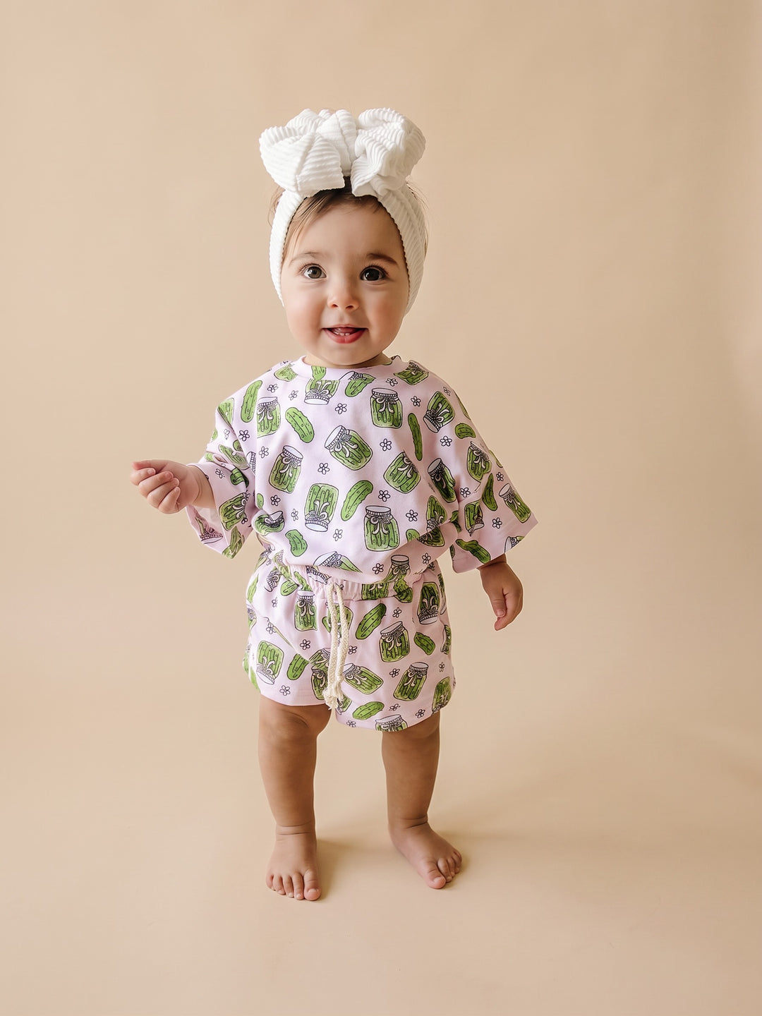 A baby girl wearing a light pink two-piece outfit with a green and gray pattern, a white bow headband, and standing barefoot against a solid beige background.