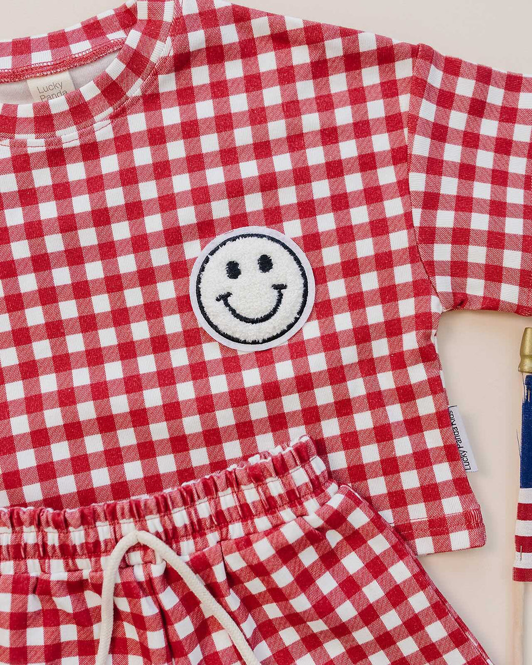 Baby wearing Lucky Panda Kids Red Gingham Shorts Set — gender neutral cotton two-piece with smiley face patch
Lucky Panda Kids Shorts Set Red Gingham flat lay — relaxed top with smiley patch and matching elastic waistband shorts
Toddler in Lucky Panda Kids Red Gingham Shorts Set — sizes 0–3M to 8–9Y, gender neutral summer outfit
Close-up of Lucky Panda Kids Red Gingham Shorts Set smiley face patch — soft 95% cotton kids two-piece
Lucky Panda Kids Red Gingham Shorts Set on baby — gender neutral cotton summer