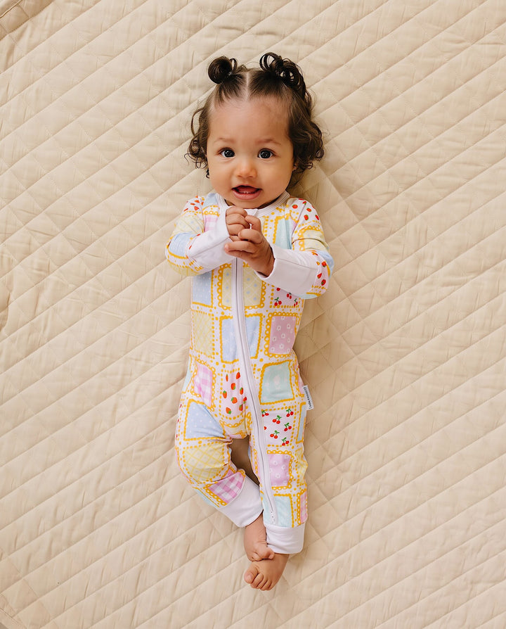 Baby lying on a cream quilt in the Sweet Patchwork zip romper with pastel printed squares, yellow borders and white cuffs.