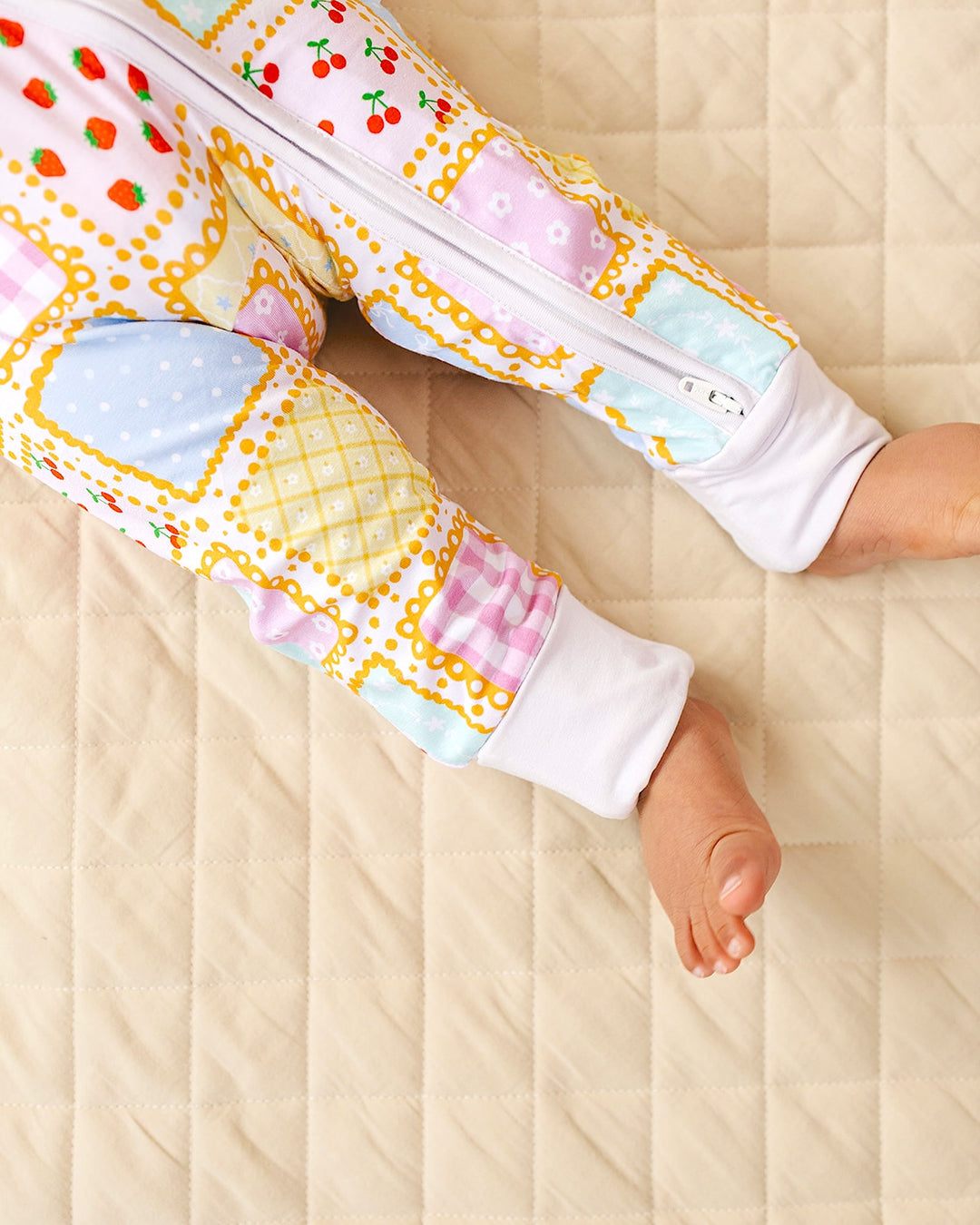 Close up of the Sweet Patchwork romper legs showing pastel squares, white ankle cuffs and uncovered feet on a quilt.