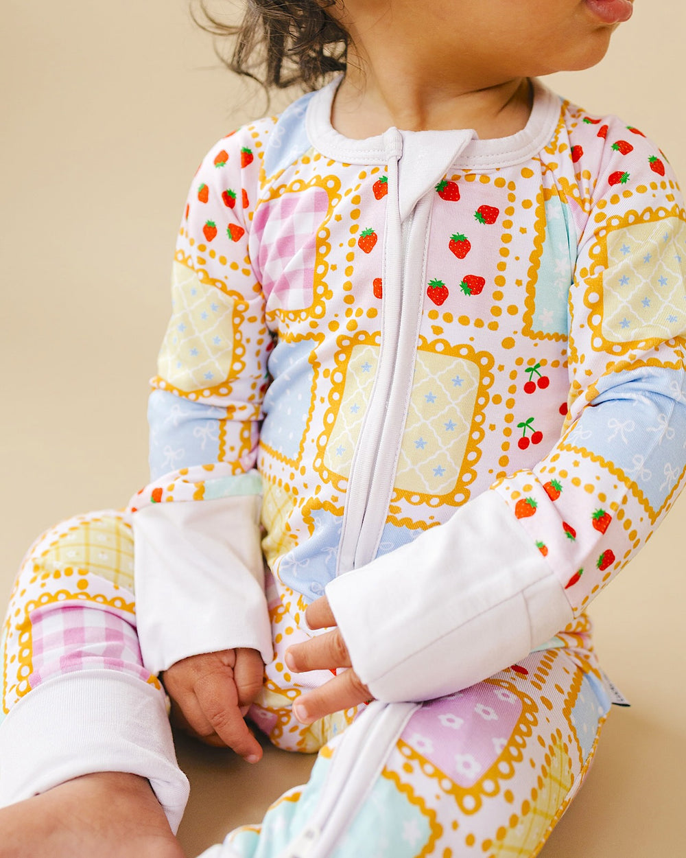 Seated baby wearing the Sweet Patchwork romper, showing the white front zipper, wrist cuffs and pastel patchwork print.