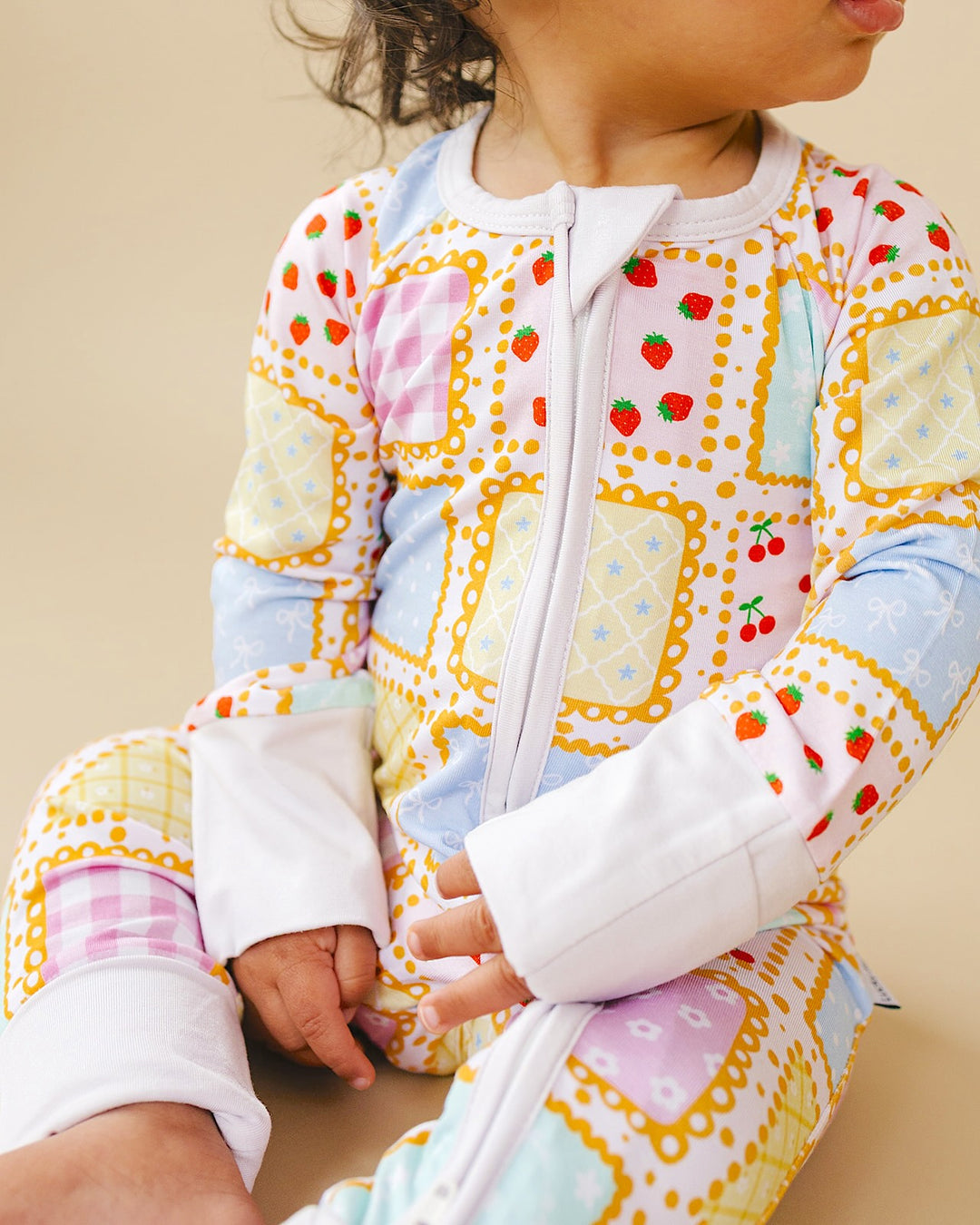 Seated baby wearing the Sweet Patchwork romper, showing the white front zipper, wrist cuffs and pastel patchwork print.