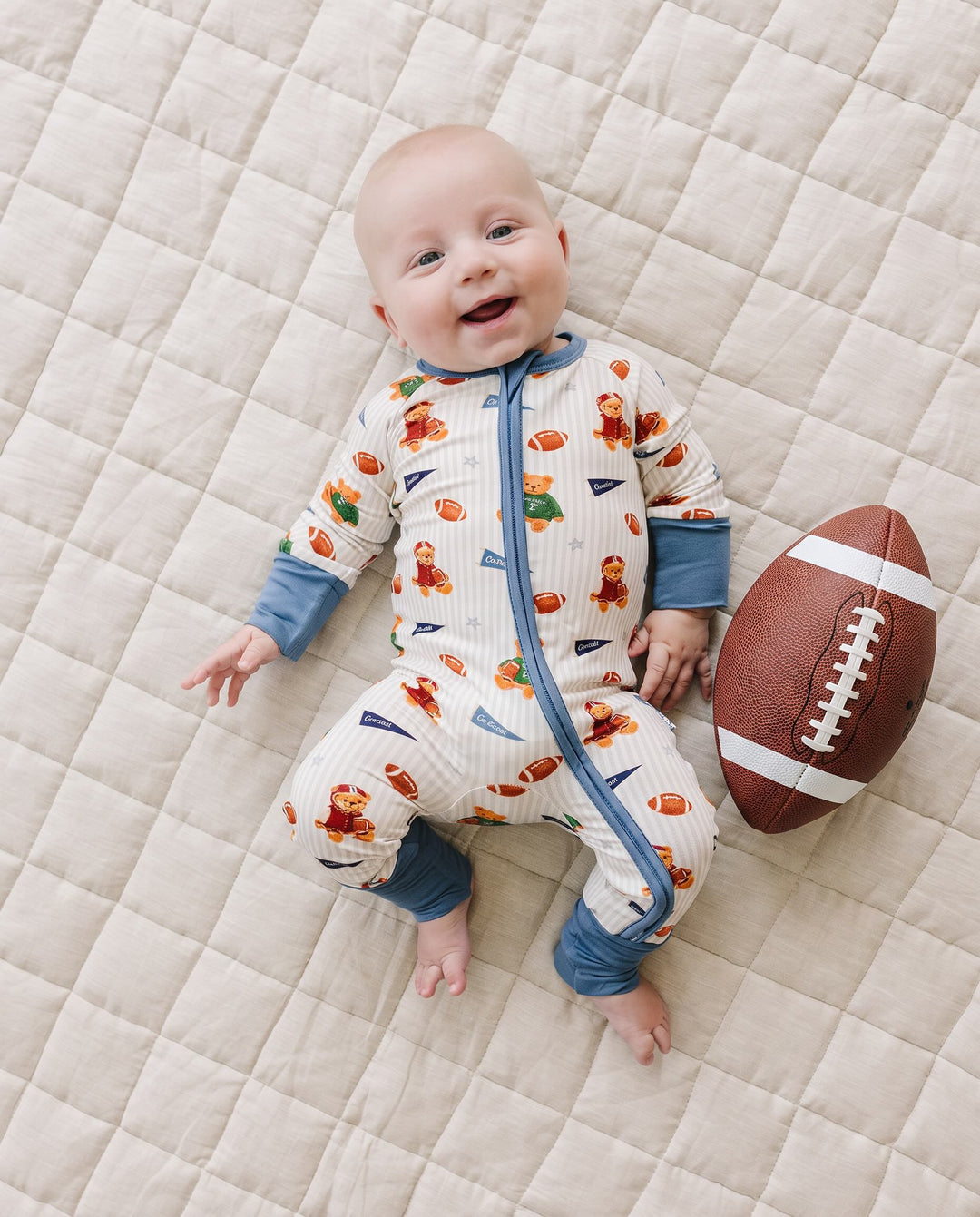 Baby wearing the Touchdown Teddy bamboo zip romper beside a football on a cream quilted mat.