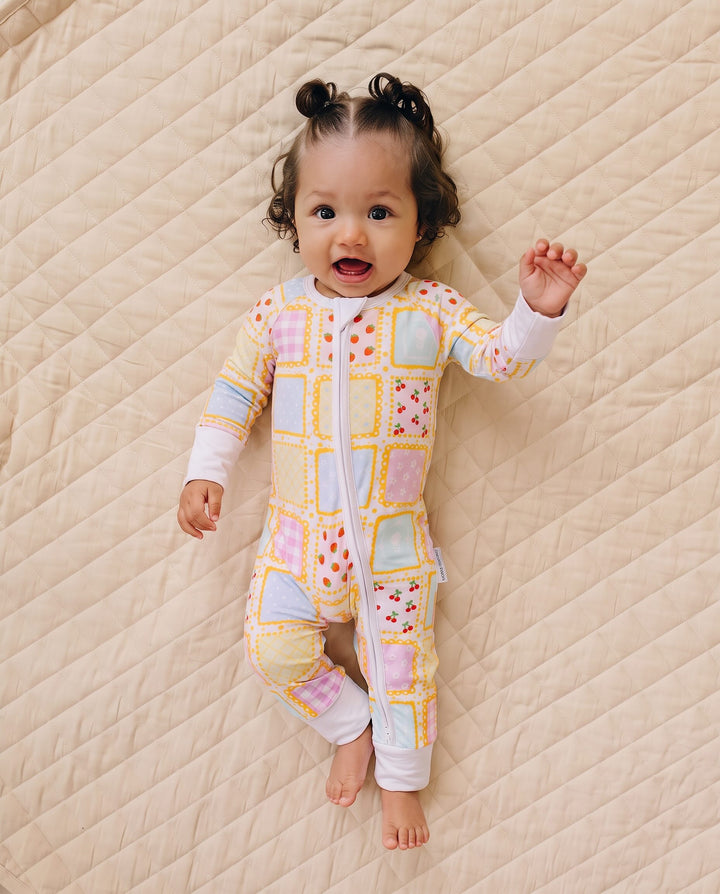 Baby smiling with one hand raised while lying on a quilt in the Sweet Patchwork zip romper with feet uncovered.
